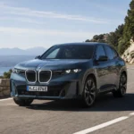 سيارة بي إم دبليو iX3 الكهربائية الجديدة أثناء القيادة على طريق جبلي ساحلي، بتصميم SUV فاخر وإضاءة LED حديثة مع خلفية طبيعية وإطلالة بحرية هادئة.