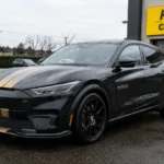 سيارة فورد موستانج Mach-E شيلبي الكهربائية باللون الأسود مع الخطوط الذهبية متوقفة أمام فرع Hertz Car Sales في أجواء ممطرة، في مشهد يعكس تخفيضات السيارات الكهربائية الرياضية النادرة.
