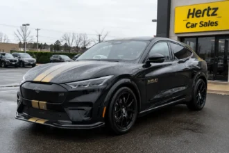 سيارة فورد موستانج Mach-E شيلبي الكهربائية باللون الأسود مع الخطوط الذهبية متوقفة أمام فرع Hertz Car Sales في أجواء ممطرة، في مشهد يعكس تخفيضات السيارات الكهربائية الرياضية النادرة.