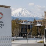 مشهد لمدينة Toyota Woven City الذكية في اليابان مع حافلة كهربائية ذاتية القيادة ومبانٍ مستقبلية حديثة أمام جبل فوجي.